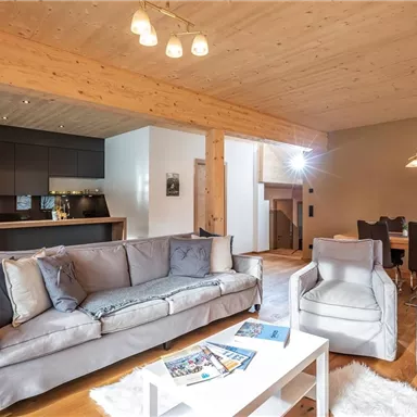 Ein modernes Wohnzimmer mit einem großen Sofa, zwei Sesseln und einem weißen Couchtisch. Im Hintergrund befindet sich eine Küchenzeile und ein Essbereich.