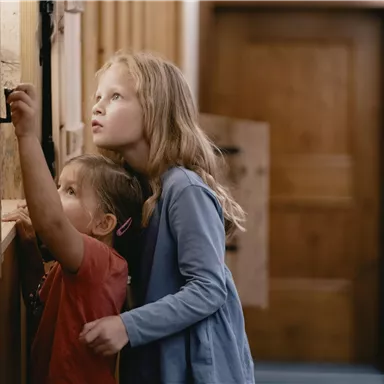 Zwei Kinder stehen in einem Flur und schauen neugierig auf eine Wand. Das Mädchen beobachtet das Geschehen, während das kleine Kind daneben steht.