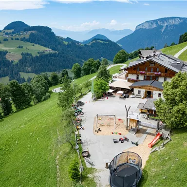 Eine malerisches Hotel umgeben von grünen Wiesen und Bergen. Im Vordergrund ist ein Spielplatz zu sehen.