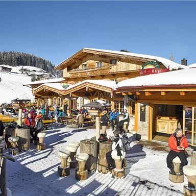 Eine gemütliche Berghütte im Schnee mit vielen Gästen. Die Sonne scheint auf die Terrasse, wo sich Menschen entspannen und die winterliche Landschaft genießen.