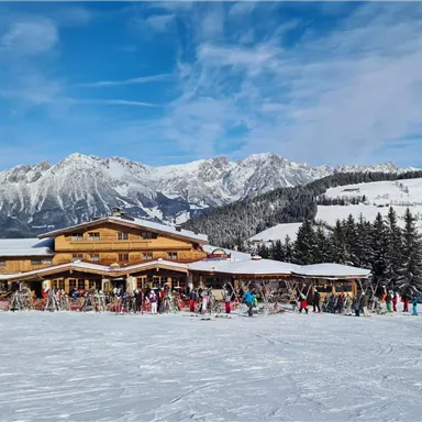 Ein gemütliches Alpengasthaus in den Bergen, umgeben von Schnee. Viele Menschen stehen vor dem Gebäude und genießen den Wintertag.