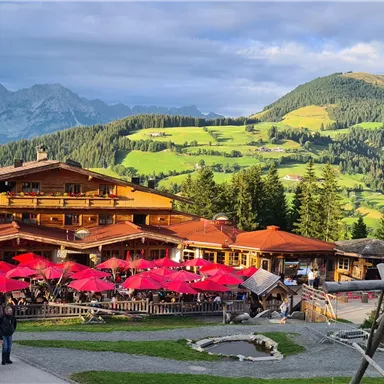 Ein gemütliches alpenländisches Gasthaus mit roten Sonnenschirmen und vielen Gästen. Im Hintergrund sind grüne Hügel und Berge zu sehen.