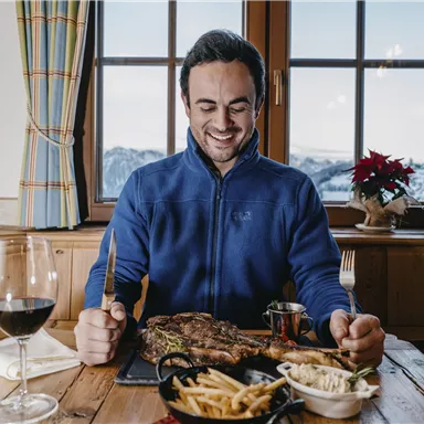 Ein Mann sitzt an einem Tisch und lächelt, während er ein großes Stück Fleisch betrachtet. Auf dem Tisch sind auch Pommes, Salat und ein Glas Rotwein zu sehen.