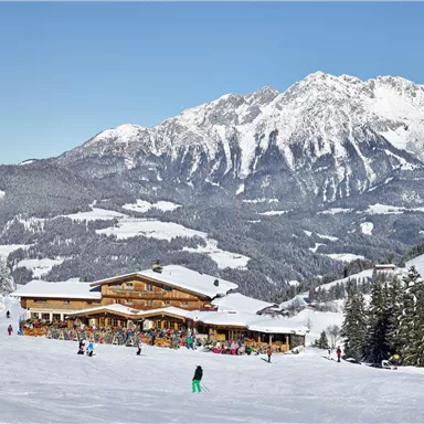 Ein malerischer Winterort mit einer gemütlichen Berghütte. Im Hintergrund ragen schneebedeckte Berge unter einem klaren blauen Himmel auf.