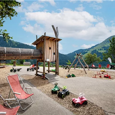 Ein Spielplatz in der Natur mit einer Holzspielstruktur und Schaukeln. Viele kleine Traktoren stehen bereit, um von Kindern genutzt zu werden.