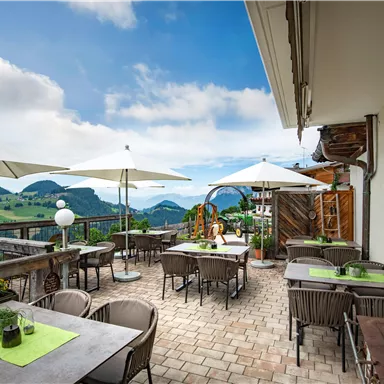 Eine gemütliche Terrasse mit Tischen und Stühlen, umgeben von schöner Landschaft. Sonnenschirme bieten Schatten, während die Aussicht auf die Berge beeindruckt.