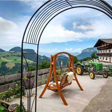 Eine gemütliche Terrasse mit einer Schaukel und Blick auf grüne Hügel und Berge. Im Hintergrund steht ein Traktor und ein traditionelles alpenländisches Haus.