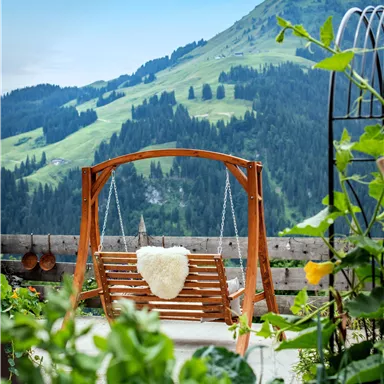 Eine Holzschaukel steht im Vordergrund, umgeben von grünen Pflanzen. Im Hintergrund erstrecken sich sanfte Hügel unter einem klaren blauen Himmel.
