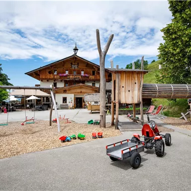 Ein Spielplatz mit verschiedenen Spielgeräten und Kettcars befindet sich vor einem rustikalen Holzgebäude. Die Umgebung ist grün mit Bäumen und einem klaren Himmel.