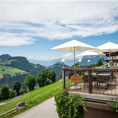 Eine gemütliche Terrasse mit Sonnenschirmen und Blick auf grüne Hügel. Die Landschaft ist von Bergen umgeben und der Himmel ist teilweise bewölkt.