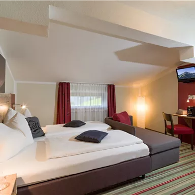 Ein gemütliches Zimmer mit zwei Betten, einem Sofa und einem Schreibtisch. Helle Farben und ein Fenster sorgen für eine freundliche Atmosphäre.