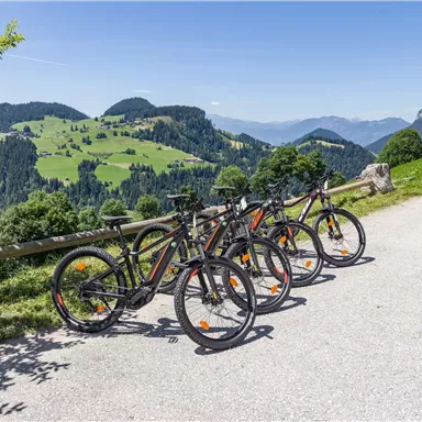 Drei Fahrräder stehen an einem Weg mit Blick auf grüne Hügel und Berge. Die Landschaft ist sonnig und einladend.