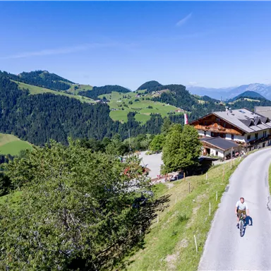 Eine malerische Landschaft mit grünen Hügeln und Bergen im Hintergrund. Auf der Straße fährt ein Radfahrer vorbei, während ein rustikales Gebäude in der Nähe steht.