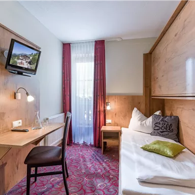 Ein gemütliches Zimmer mit Holzmöbeln und einem Fernseher an der Wand. Es gibt einen kleinen Tisch, einen Stuhl und ein bequemes Bett mit Dekokissen.