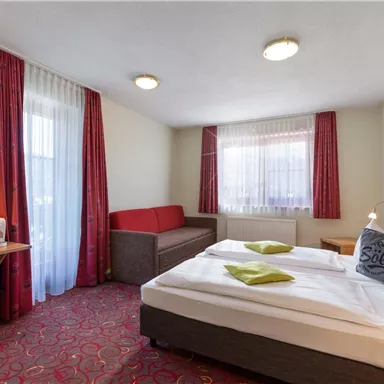 Ein gemütliches Hotelzimmer mit zwei Betten, einem Sofa und einem kleinen Tisch. Die Fenster sind mit Vorhängen dekoriert und der Boden hat einen roten Teppich.
