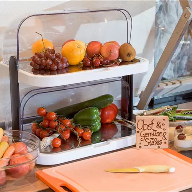 Eine Ablage mit frischem Obst und Gemüse, darunter Äpfel, Trauben, Tomaten und Gurken. Daneben steht ein Holzschild mit der Aufschrift "Obst & Gemüse & Säfte".