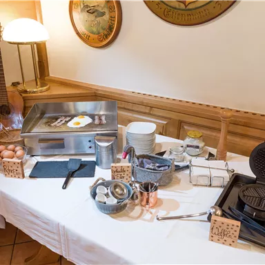 Ein Frühstücksbuffet mit einer Grillplatte, Eiern und verschiedenen Kochutensilien. Im Hintergrund steht eine Gitarre, was eine gemütliche Atmosphäre schafft.