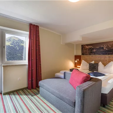 Ein gemütliches Hotelzimmer mit einem großen Fenster, das Ausblick auf die Berge bietet. Die Einrichtung ist modern mit einem Doppelbett und einem bequemen Sofa.