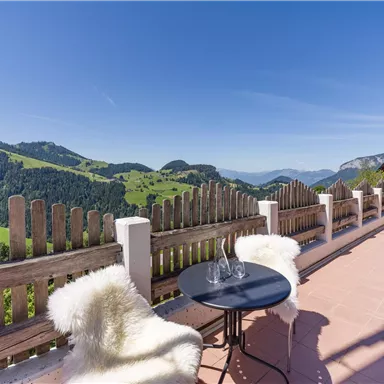 Eine sonnige Terrasse mit Blick auf grüne Hügel und Berge. Ein Tisch mit zwei Stühlen lädt zum Entspannen ein.