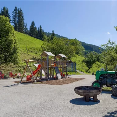 Ein Spielplatz mit Rutschen und Schaukeln, umgeben von grünen Wiesen. Im Hintergrund sind Berge und ein alter Traktor zu sehen.