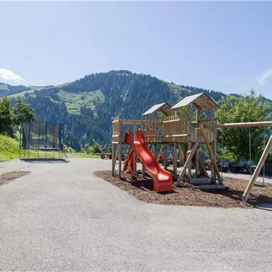 Ein Spielplatz mit einer Rutsche und einem Klettergerüst in einer malerischen Berglandschaft. Im Hintergrund sind grüne Hügel und ein blauer Himmel zu sehen.