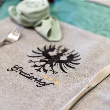 Ein gedeckter Tisch mit einem grauen Platzdeckchen, auf dem ein Wappen abgebildet ist. Besteck und ein dekoriertes Serviettenröllchen ergänzen das Arrangement.