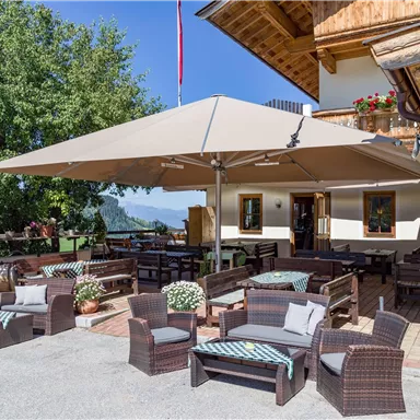 Eine gemütliche Terrasse mit Rattanmöbeln und einem großen Sonnenschirm. Im Hintergrund sind Holzstrukturen und Bäume zu sehen.