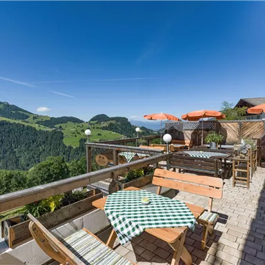 Eine gemütliche Terrasse mit Holztischen und -stühlen, umgeben von grünen Hügeln. Im Hintergrund ist ein klarer blauer Himmel zu sehen.
