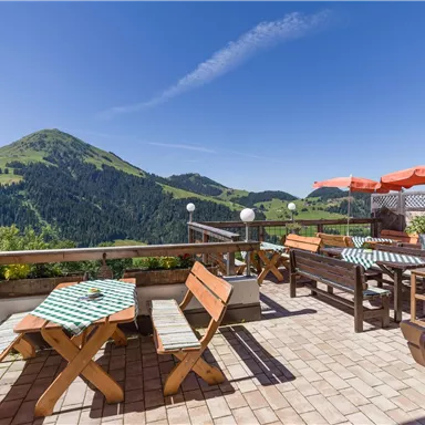 Eine sonnige Terrasse mit Holzstühlen und Tischen, umgeben von einer malerischen Berglandschaft. Im Hintergrund sind grüne Hügel und ein klarer blauer Himmel zu sehen.
