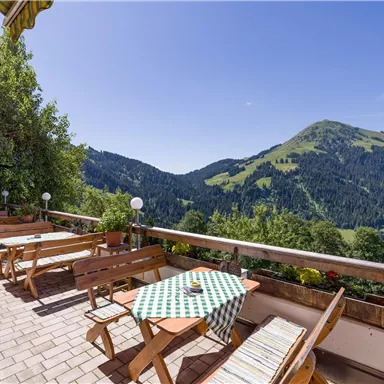 Eine gemütliche Terrasse mit Holztischen und Blick auf grüne Berge. Der klare Himmel und die umliegende Natur sorgen für eine entspannte Atmosphäre.