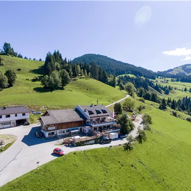 Ein malerisches, hügeliges Landschaftsbild mit grünen Wiesen und dichten Wäldern. Im Vordergrund befindet sich ein gemütliches Gasthaus mit ein paar Autos.
