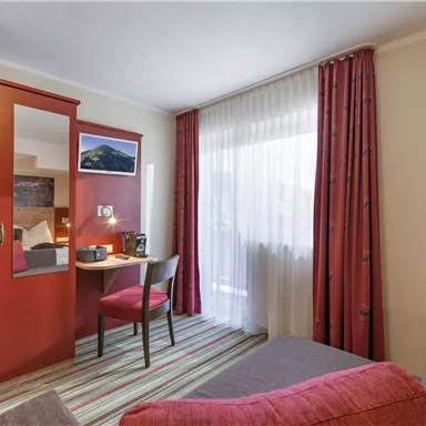 Ein gemütliches Hotelzimmer mit einem Schreibtisch, einem Stuhl und einem Kleiderschrank. Große Fenster lassen viel Licht herein und die dekorativen Vorhänge verleihen dem Raum eine einladende Atmosphäre.