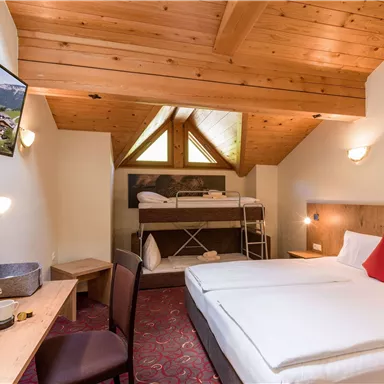 Ein gemütliches Hotelzimmer mit Holzboden und einem Doppelbett. Im Hintergrund befindet sich ein Etagenbett und ein Fernseher an der Wand.