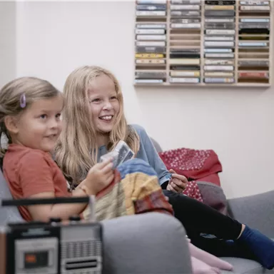 Zwei Mädchen sitzen auf einem grauen Sofa und lächeln. Im Hintergrund sind Kassetten und ein Radio sichtbar.