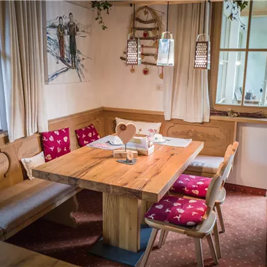 Ein gemütlicher Essbereich mit einem großen Holztisch und komfortablen Bänken. Die Wände sind freundlich dekoriert, und es gibt Fenster mit sanften Vorhängen.