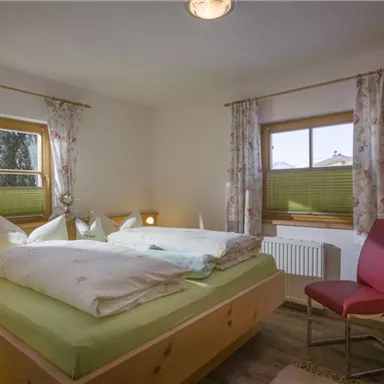 A cozy bedroom with a large double bed and two windows. The walls are light and the windows are decorated with floral curtains.