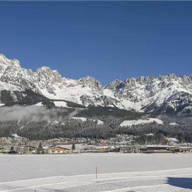 An impressive mountain landscape with snow-covered peaks and a clear sky. In the foreground, a snow-covered area extends with a small village.