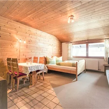 A cozy living space with a wooden ceiling and large windows. The room includes a large bed, a seating area, and a small dining table.