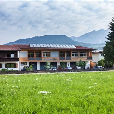 Ein modernes Gebäude umgeben von Wiesen und Bäumen in einer bergigen Landschaft. Die Wolken am Himmel verleihen der Szenerie eine ruhige Atmosphäre.