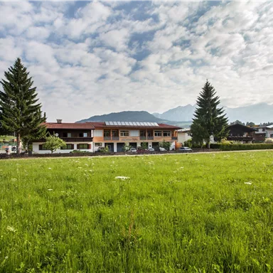 Ein malerisches Gebäude inmitten einer grünen Wiese und unter einem wolkigen Himmel. Im Hintergrund sind Berge zu sehen, die die Landschaft umrahmen.