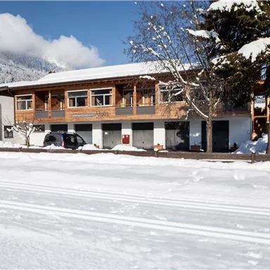 Ein winterliches Landschaftsbild mit einem Holzgebäude und schneebedecktem Boden. Im Hintergrund sind Berge unter einem blauen Himmel sichtbar.