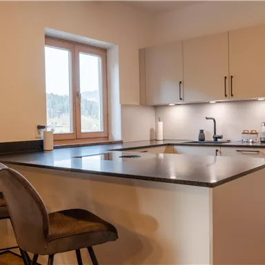 Eine moderne Küche mit einer zentralen Kochinsel und hohen Stühlen. Viel Tageslicht fällt durch das Fenster und sorgt für eine helle Atmosphäre.