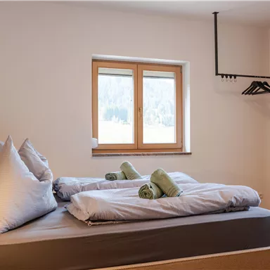 Ein helles Schlafzimmer mit einem Doppelbett und zwei grünen Handtüchern. Durch das Fenster fällt Tageslicht auf die minimalistische Ausstattung.