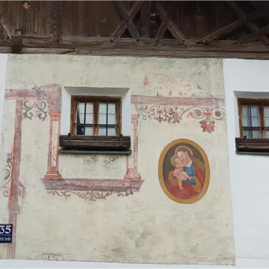 A historic building with two windows and a wall painting. The painting depicts the Madonna surrounded by decorative elements.
