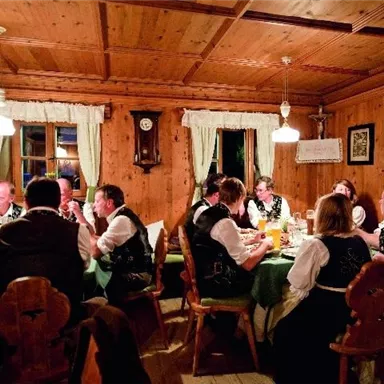 Eine gemütliche Stube mit einem langen Tisch, an dem Menschen in traditionellen Trachten zusammen essen und feiern. Warmes Licht und holzverkleidete Wände schaffen eine einladende Atmosphäre.