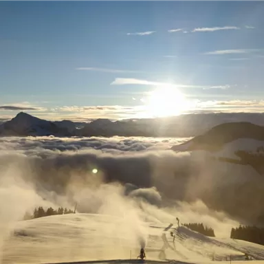 Eine atemberaubende Winterlandschaft mit schneebedeckten Bergen und einer strahlenden Sonne. Der Himmel ist klar und der Nebel umhüllt die Täler.