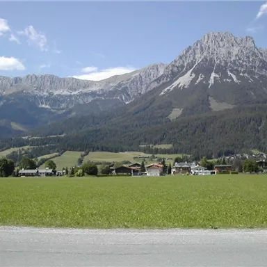 Eine malerische Landschaft mit hohen Bergen und grünen Feldern. Im Vordergrund befinden sich einige Häuser unter einem klaren blauen Himmel.