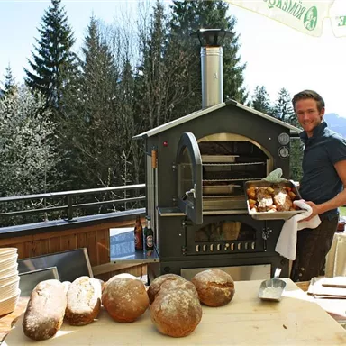 Ein Mann steht neben einem Holzofen und hält frisch gebackenes Brot in der Hand. Im Hintergrund sind hohe Bäume und ein klarer Himmel zu sehen.