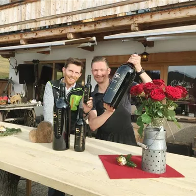 Zwei Männer stehen an einem Holztisch und halten Weinflaschen in der Hand. Im Hintergrund sind Rosen und eine gemütliche Umgebung zu sehen.