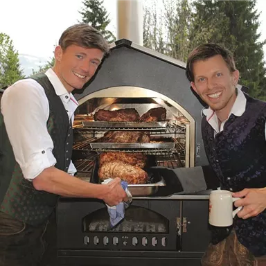 Zwei Männer in traditioneller Kleidung stehen an einem Grillofen und zeigen auf frisch gegrilltes Fleisch. Die Umgebung ist natürlich, mit Bäumen im Hintergrund.